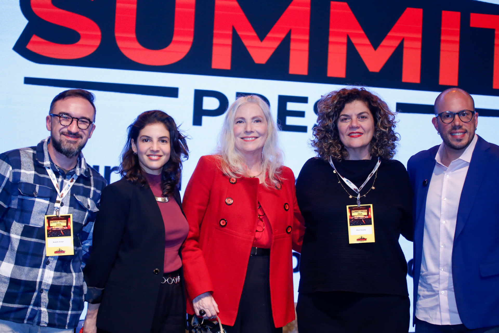 Cinco participantes do Summit Press 2025 posando diante do painel com o logotipo do evento